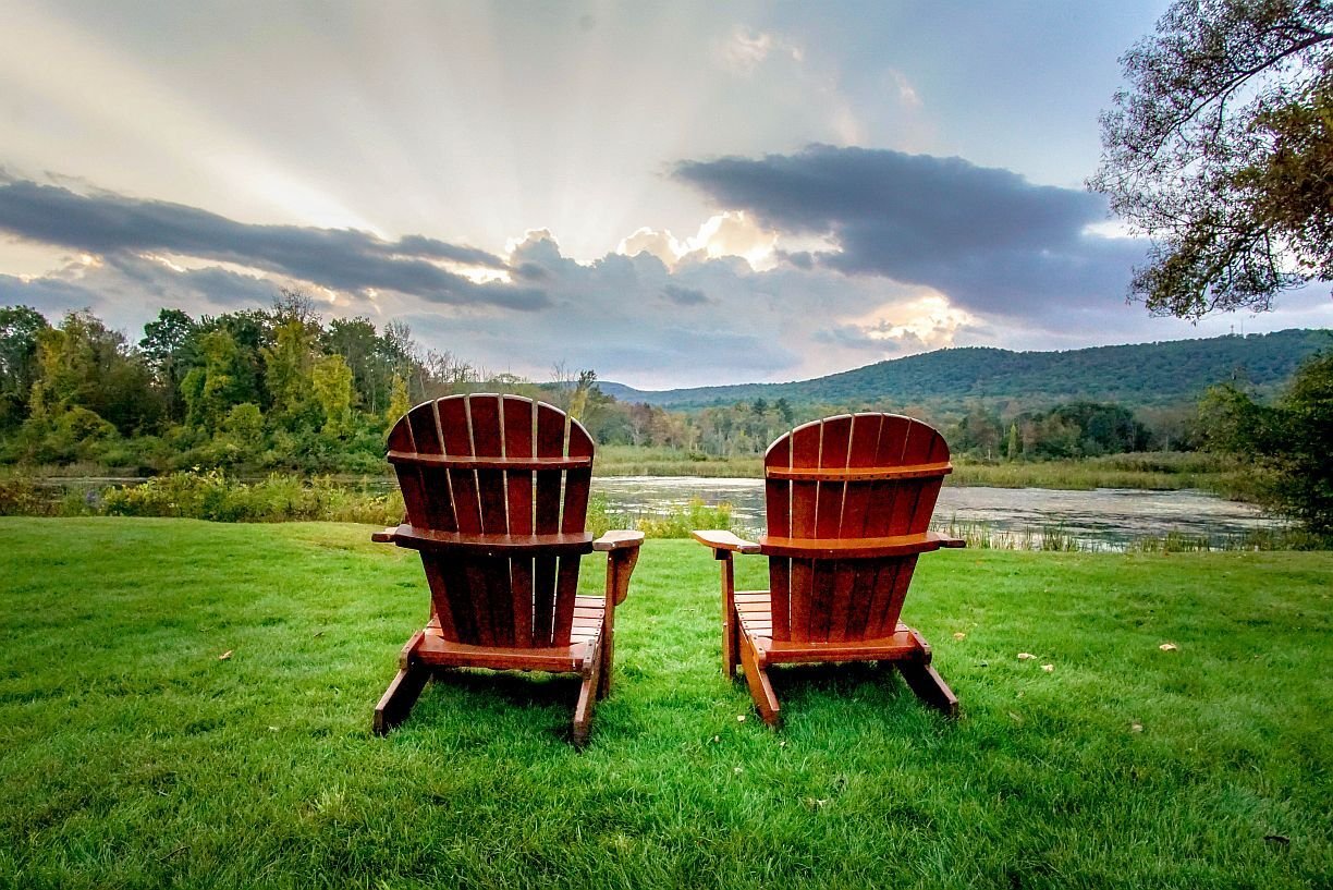 Two wooden Adirondack chairs facing a tranquil pond and mountain view at sunset on the property of The Yankee Inn